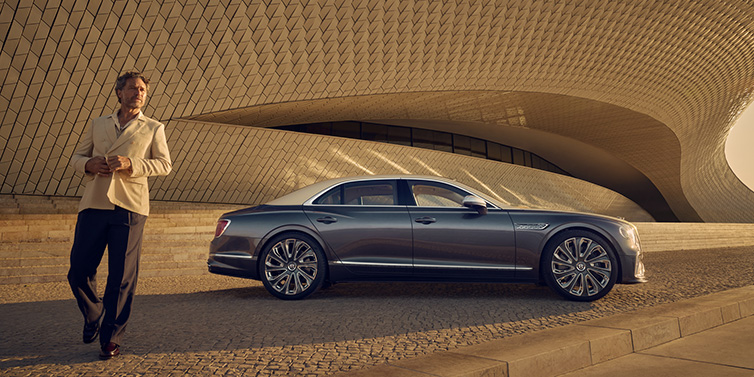 Bentley Phnom Penh Bentley Flying Spur Mulliner sedan in Rose Gold over Magnetic duotone paint parked by a curved wall with person in suit jacket standing on the left